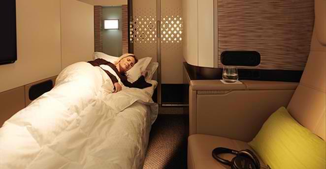 A woman sleeps comfortably in a private bed within a luxurious airplane suite, with a seat and amenities visible nearby.