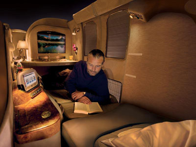 A man reads a book in a private, well-appointed airline cabin with a bed and entertainment system.