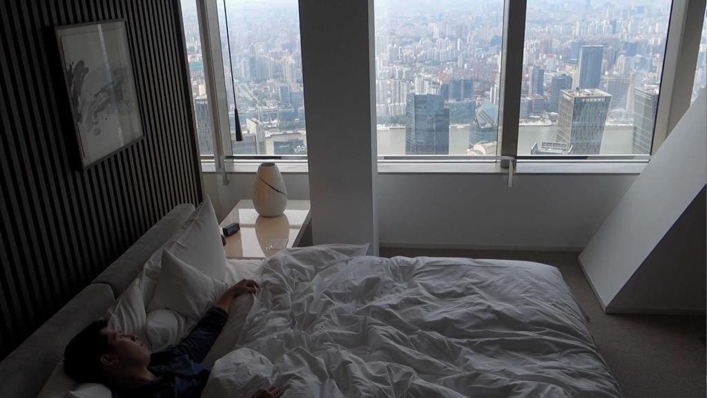 A person sleeps in a modern hotel room with a panoramic city view from large windows.