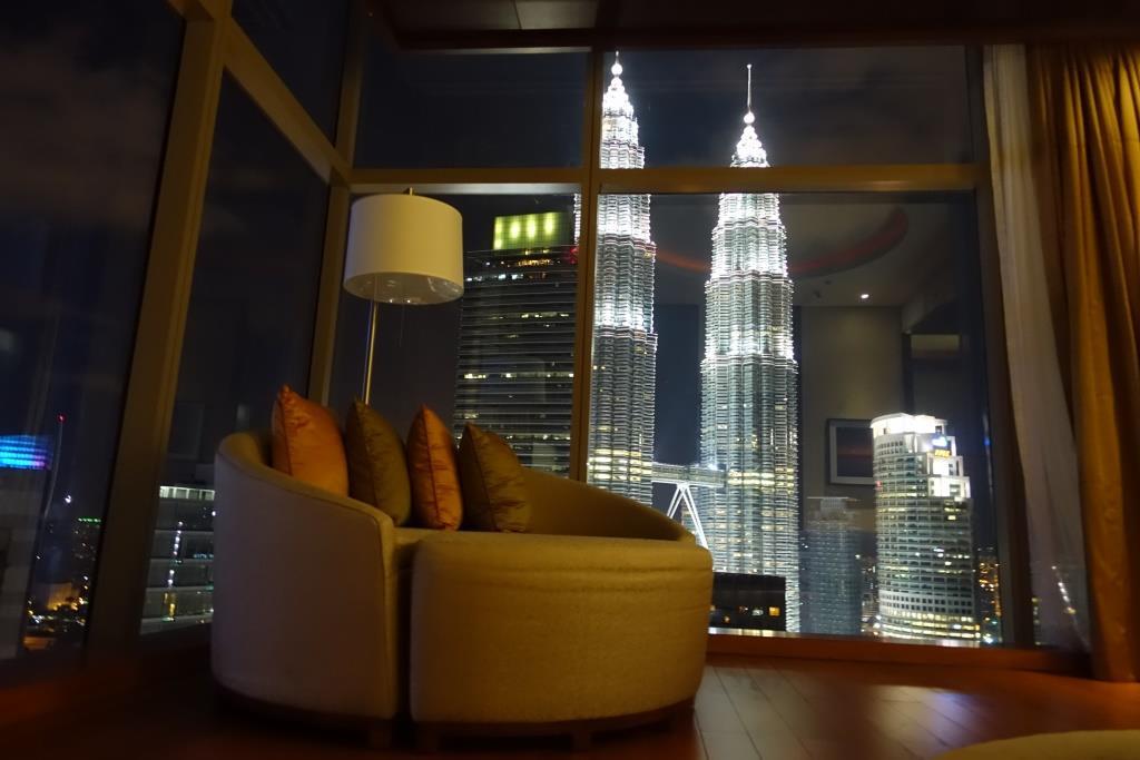 A hotel suite with a couch and lamp, overlooking the brightly lit Petronas Twin Towers at night.