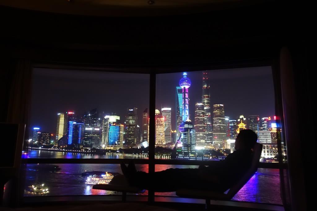A silhouetted person looks out a large window at the illuminated Shanghai skyline and river at night.