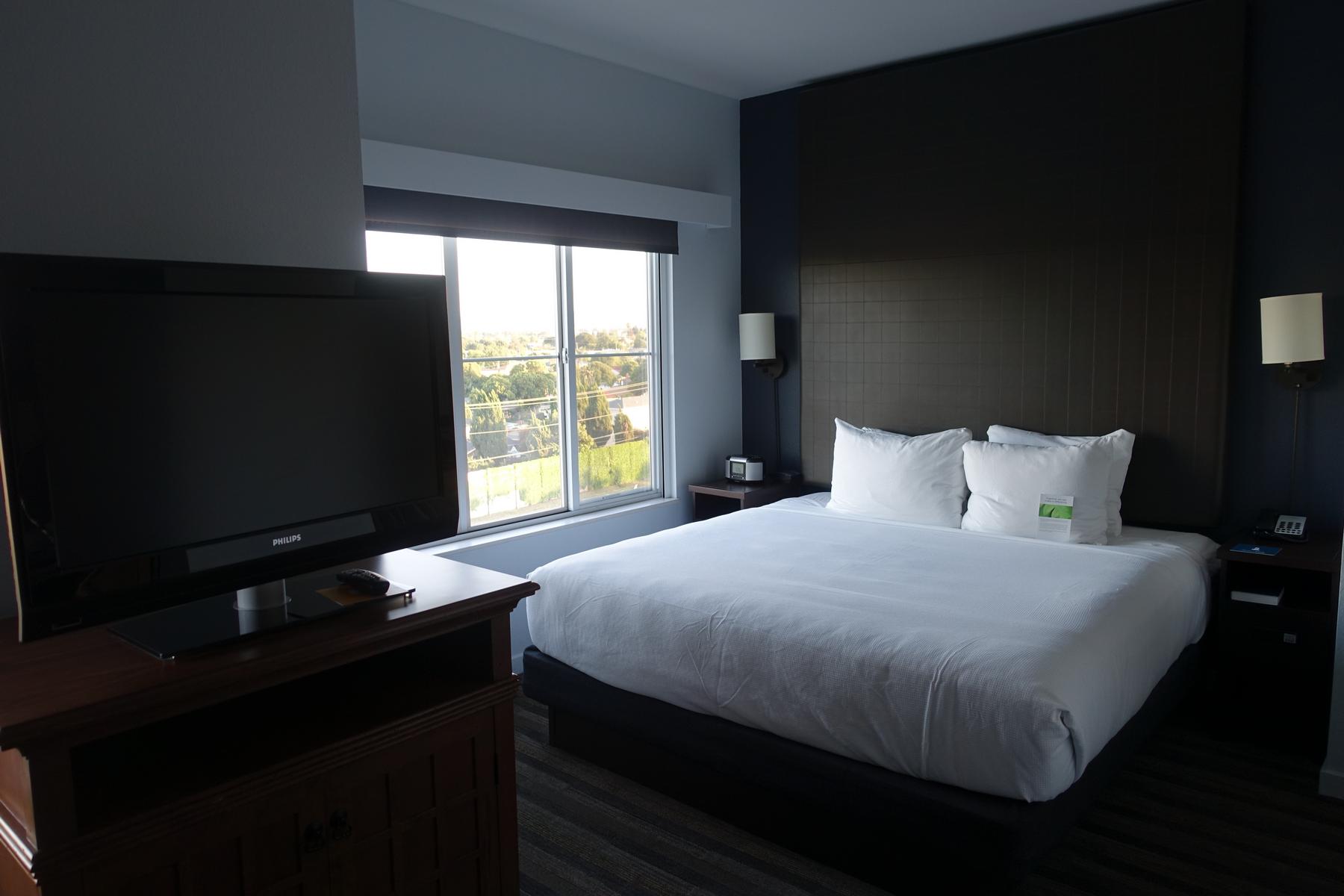 A modern hotel room with a large bed, flat-screen TV, and a window showing an outdoor view.