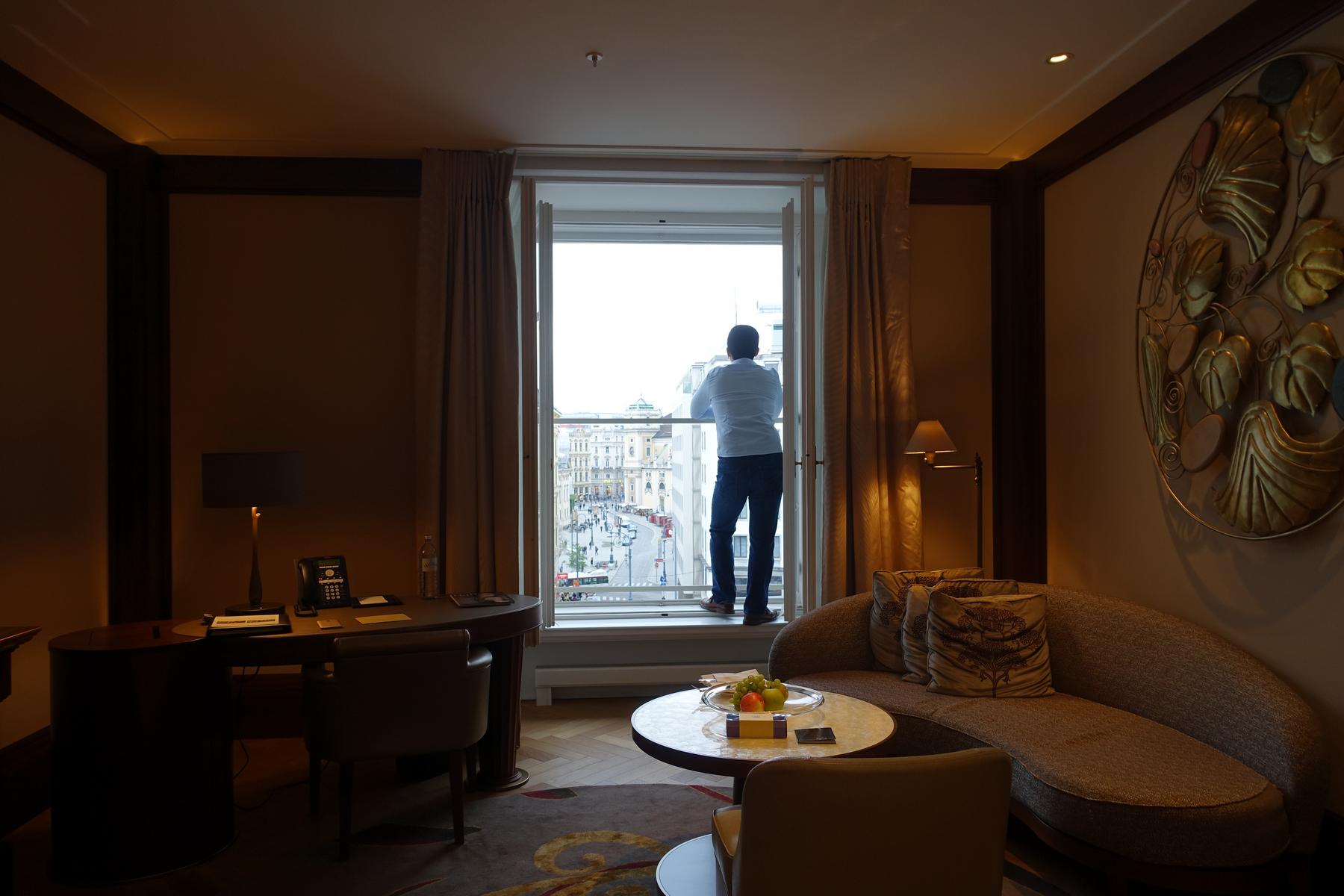 Man looking out a large window from a luxurious hotel room overlooking a city street.
