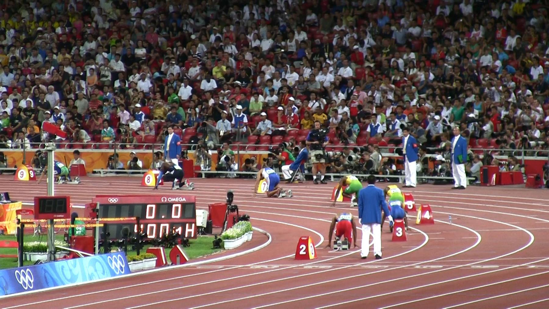 Sprinters crouch at the starting blocks of the 100m final at the Beijing 2008 Olympic Games, watched by a large crowd.