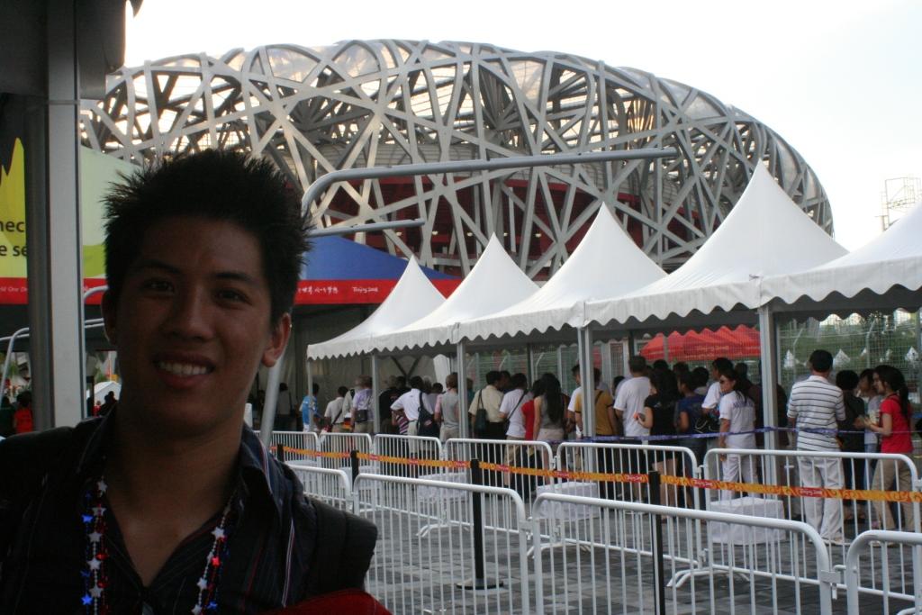 A smiling man stands in front of Beijing's Bird's Nest stadium, with crowds and white tents visible, during the 2008 Olympic Games.