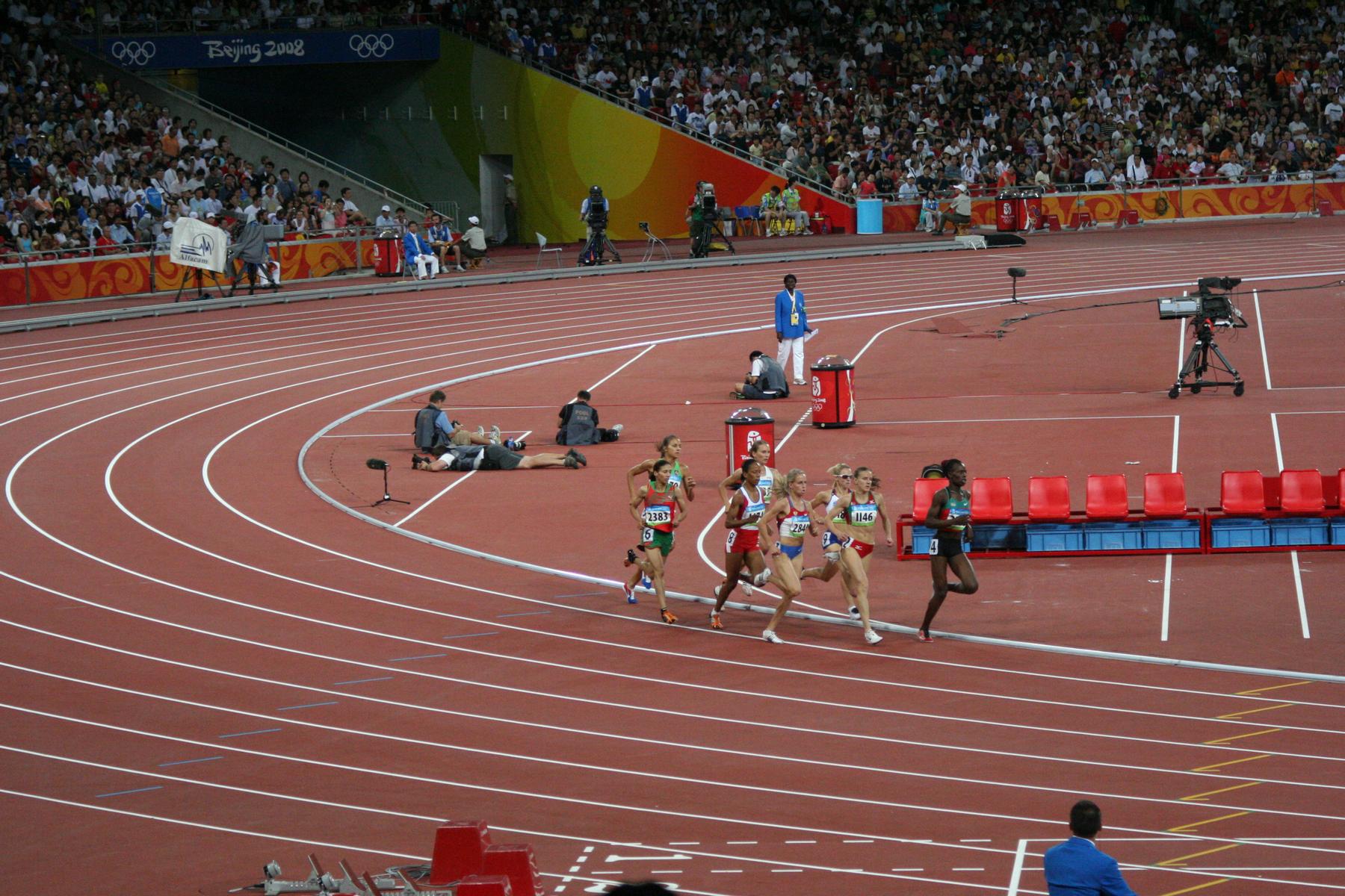 Female athletes race on a track at the Beijing 2008 Olympics stadium, with spectators in the background.