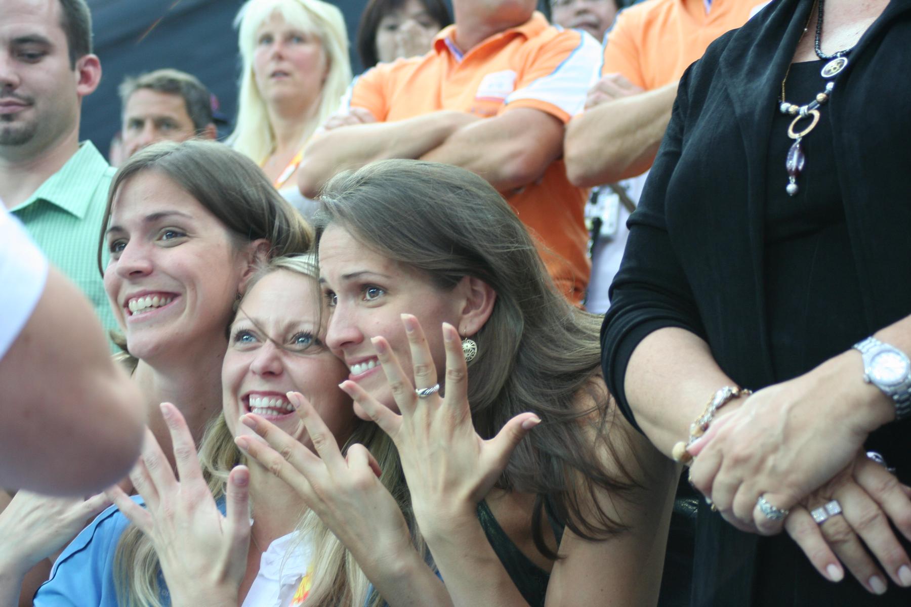 Three excited women in the foreground smile widely with raised hands, watching an event with other spectators.