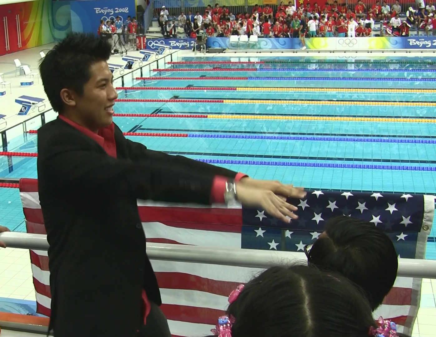 An enthusiastic man watches the Beijing 2008 Olympic swimming competition, with an American flag draped on the railing.