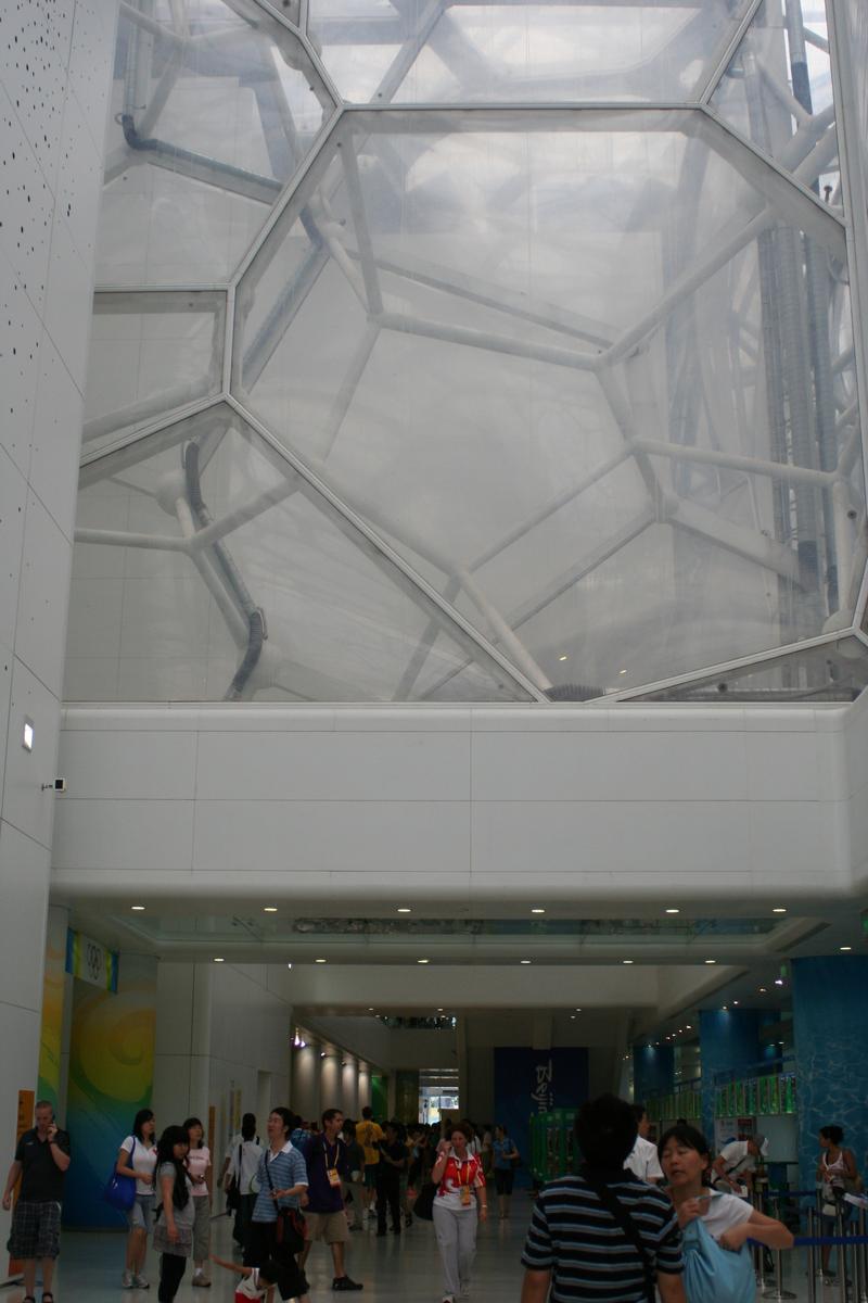 Inside the Beijing Water Cube, a geometric bubble-like ceiling looms over a wide hallway filled with people.
