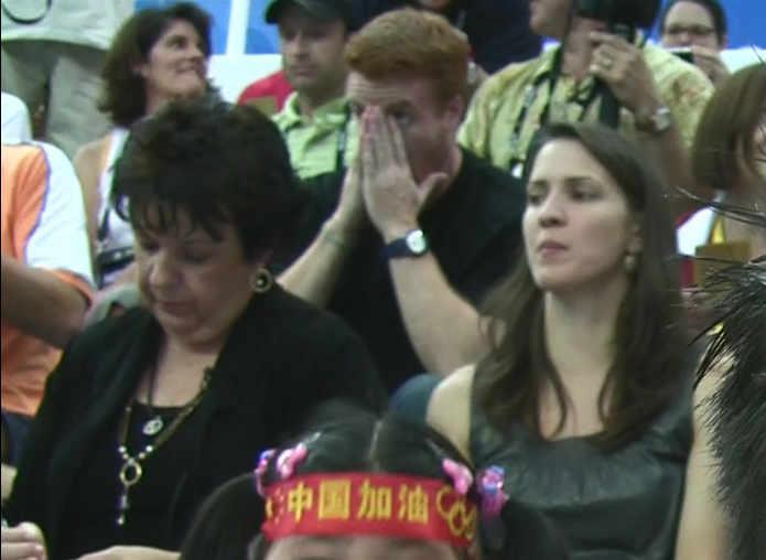 Spectators, including a man covering his face in emotion, watch an Olympic event.