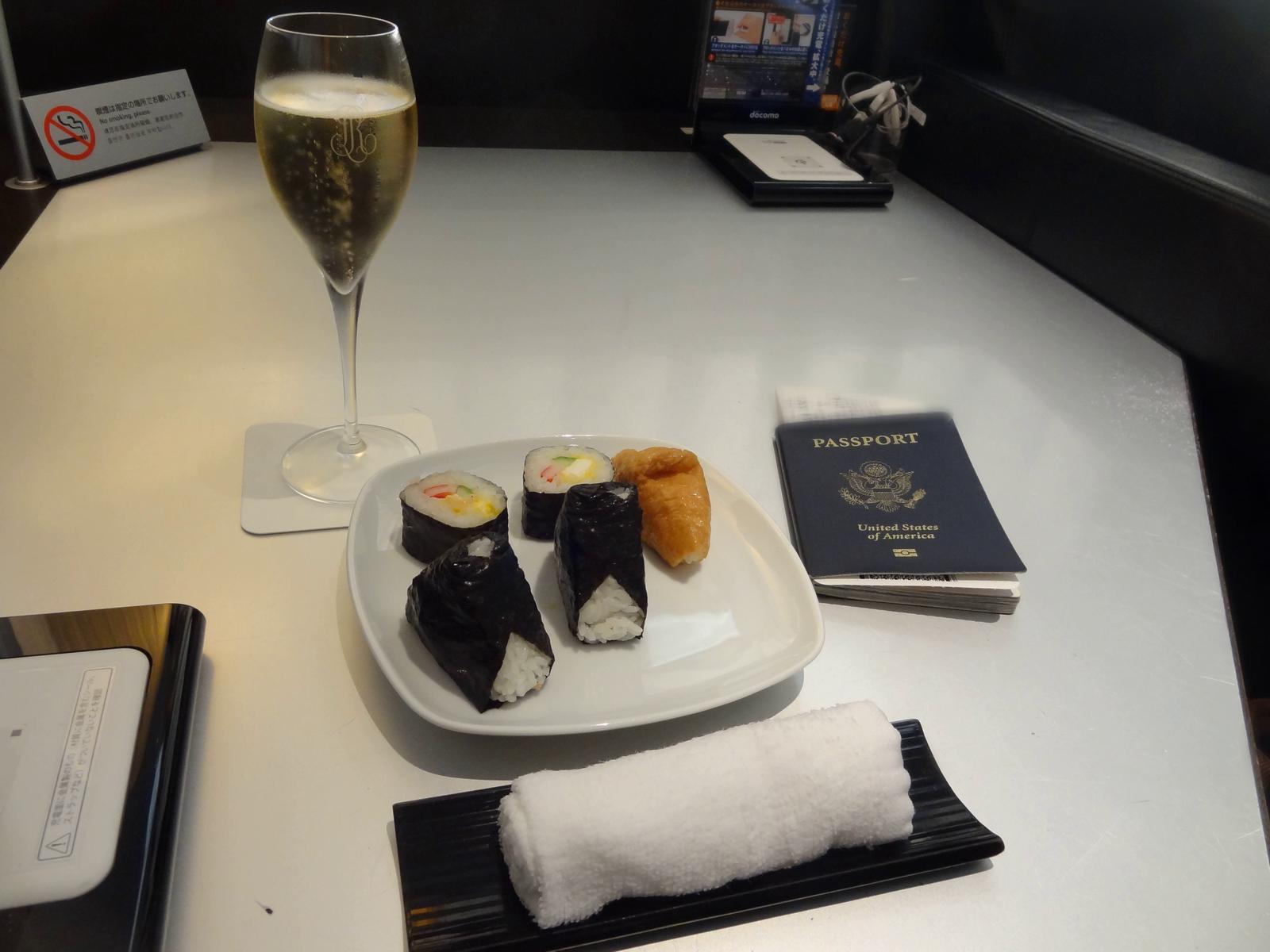 A glass of champagne, a plate of sushi, a rolled white towel, and a U.S. passport are on a table.