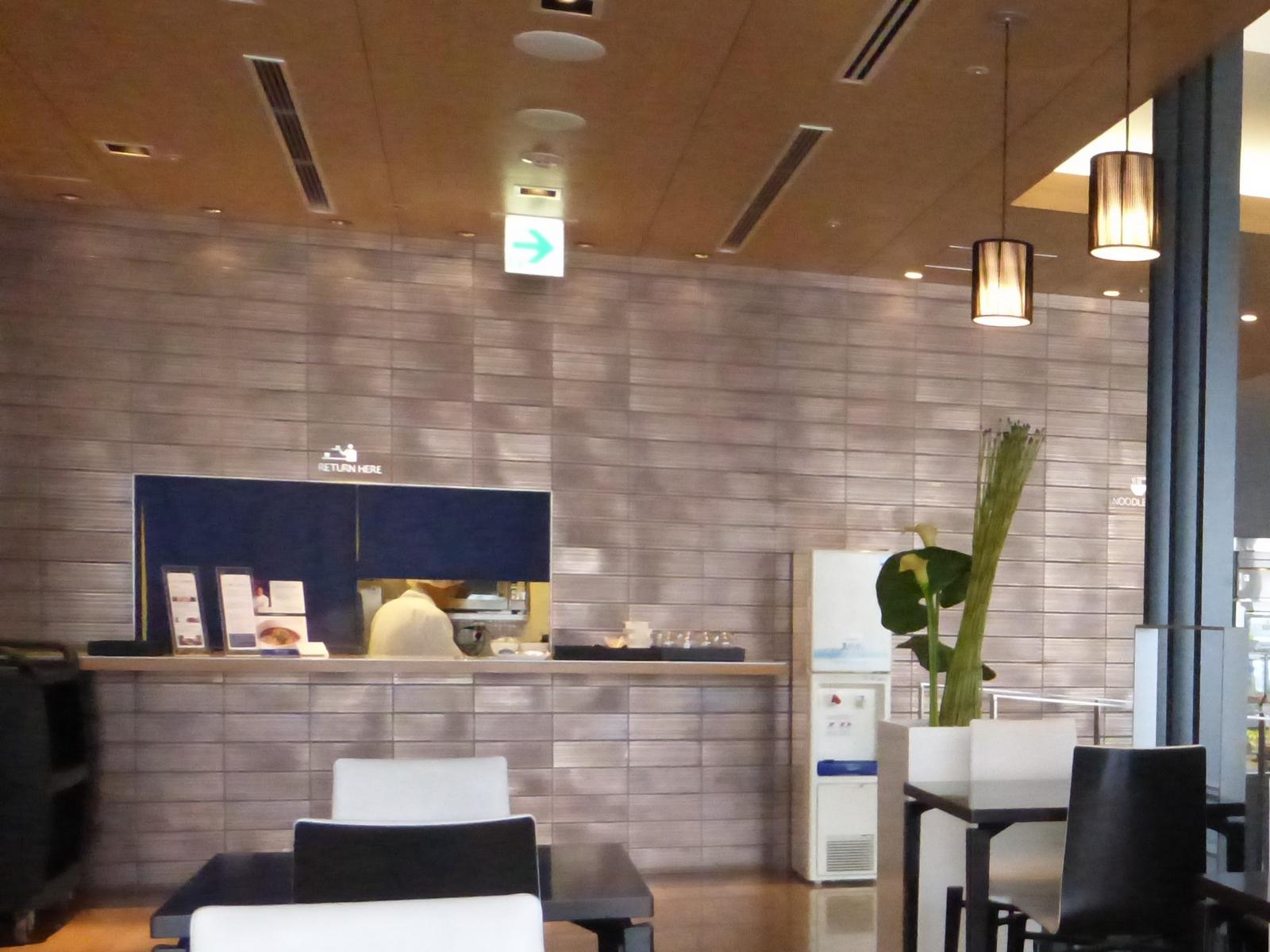 A modern dining area in the ANA First Class Lounge featuring a noodle bar counter, a server, and dining tables.