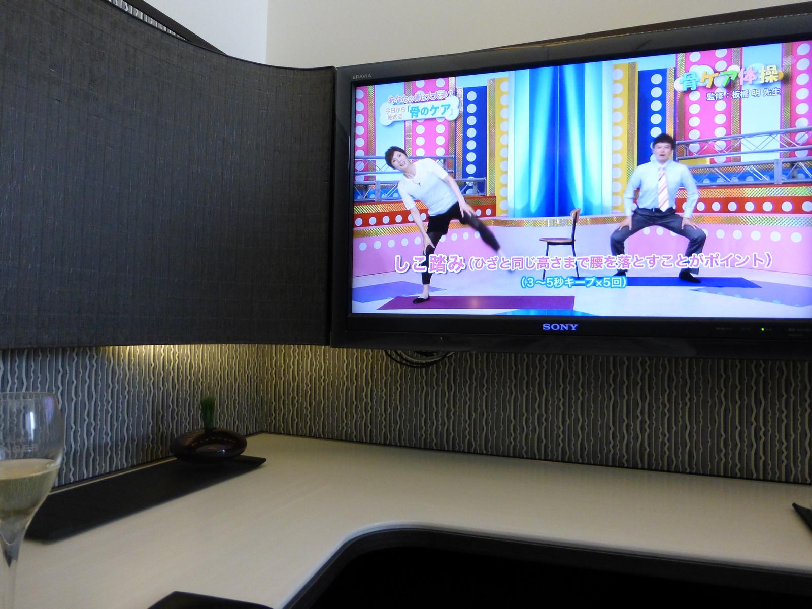 A lounge corner with a TV showing an exercise video and a glass of champagne on a desk.