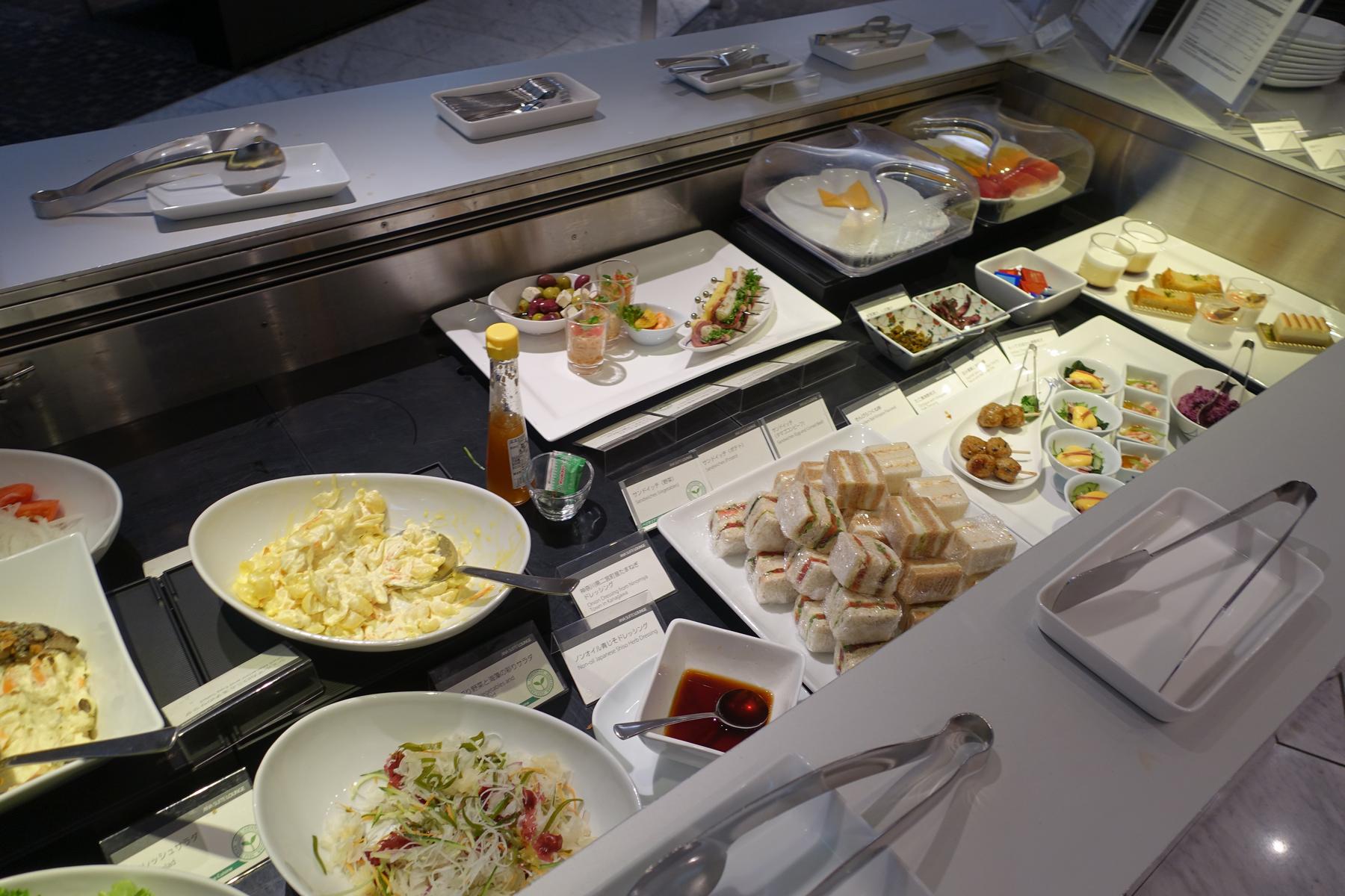 A buffet spread at the ANA First Class Lounge featuring sandwiches, salads, various appetizers, and desserts.