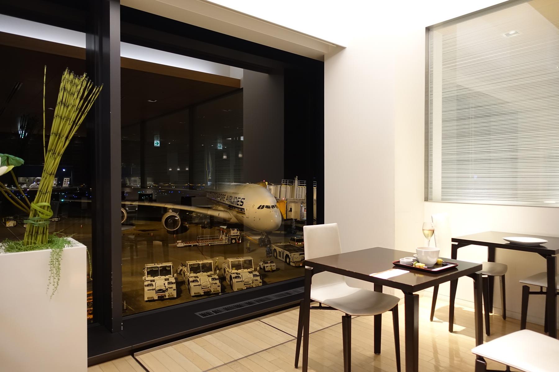 Inside an ANA First Class Lounge, a dining area looks out onto a Singapore Airlines plane parked at the gate at night.