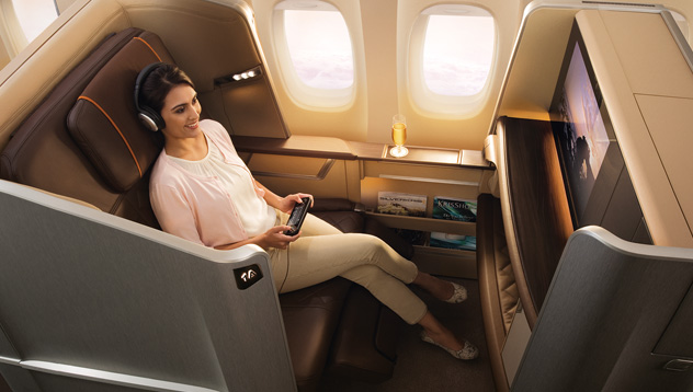 A smiling woman in headphones relaxes in a luxurious Singapore Airlines First Class Suite, holding a remote control.