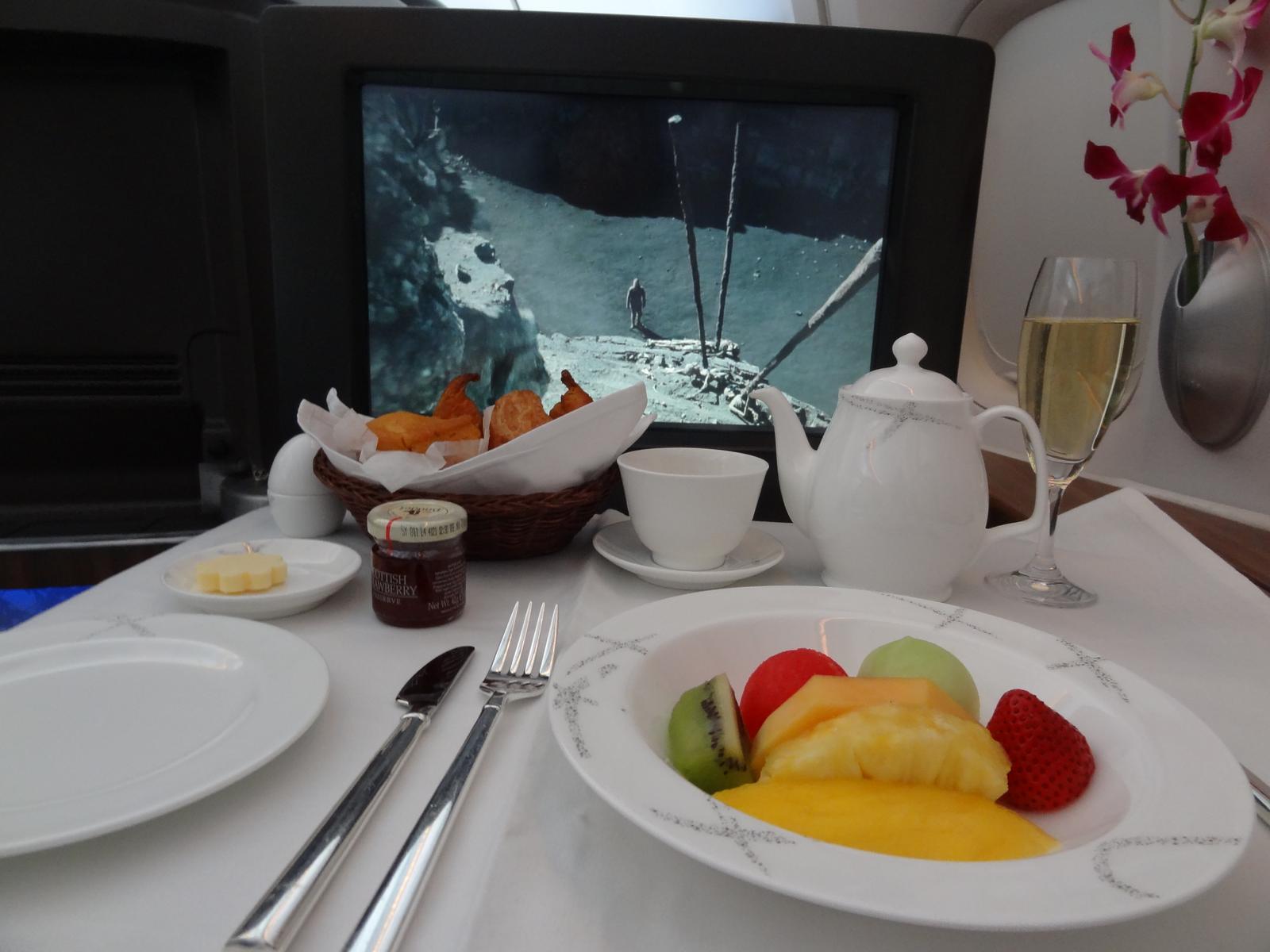 Cathay Pacific First Class breakfast setup with fruit, pastries, tea, champagne, and a movie playing on a personal screen.