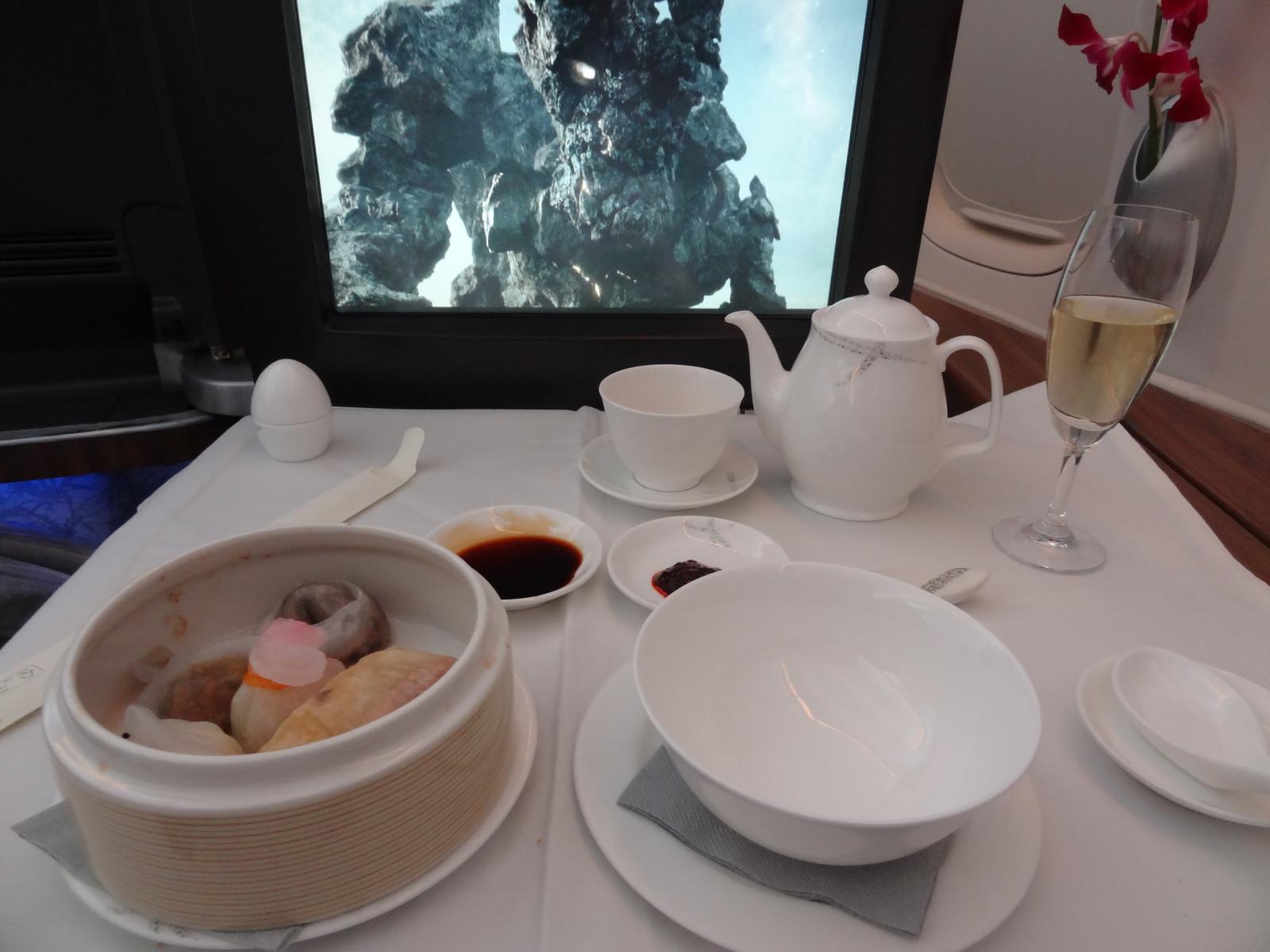 Cathay Pacific First Class meal with dim sum, tea, champagne, and a movie playing on the screen.