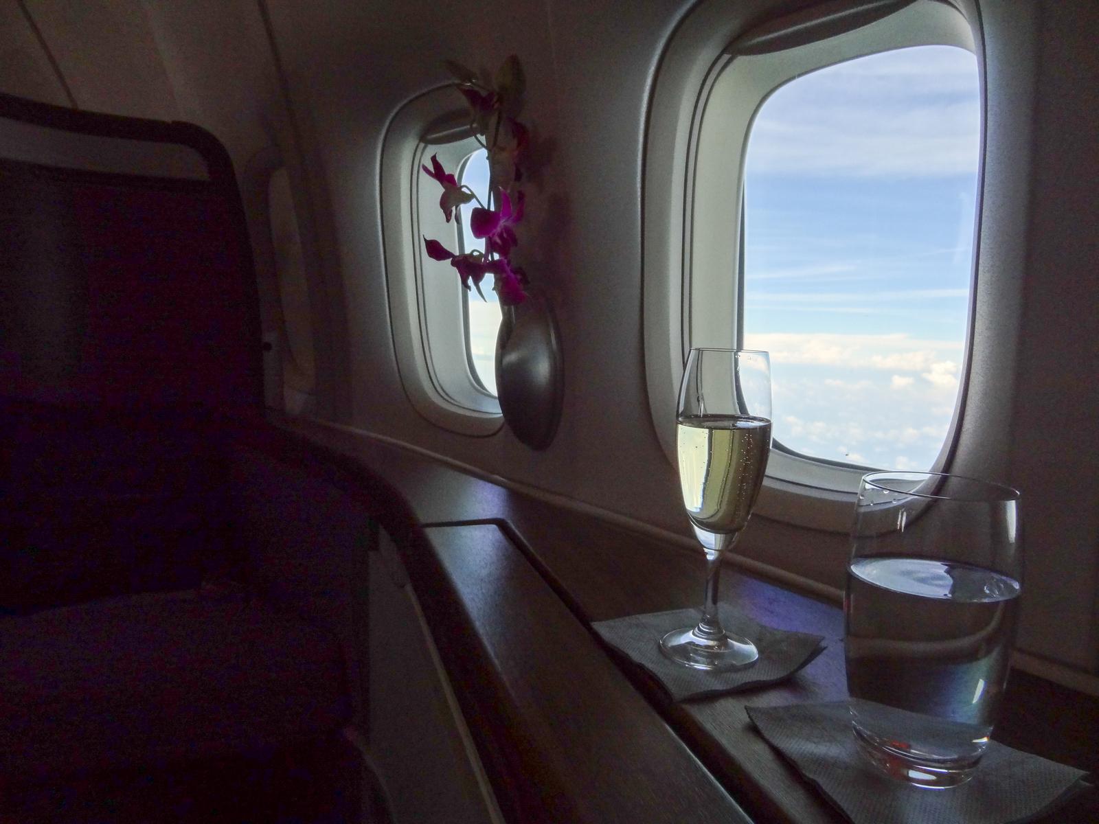 Champagne, water, and orchids on a ledge by first class airplane windows overlooking clouds.