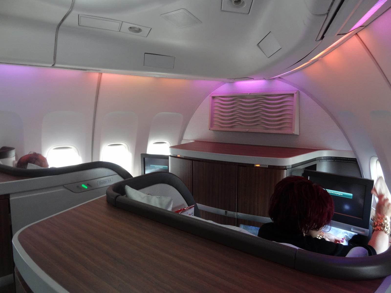 A passenger sits in a luxurious Cathay Pacific First Class suite with ambient purple and orange lighting.