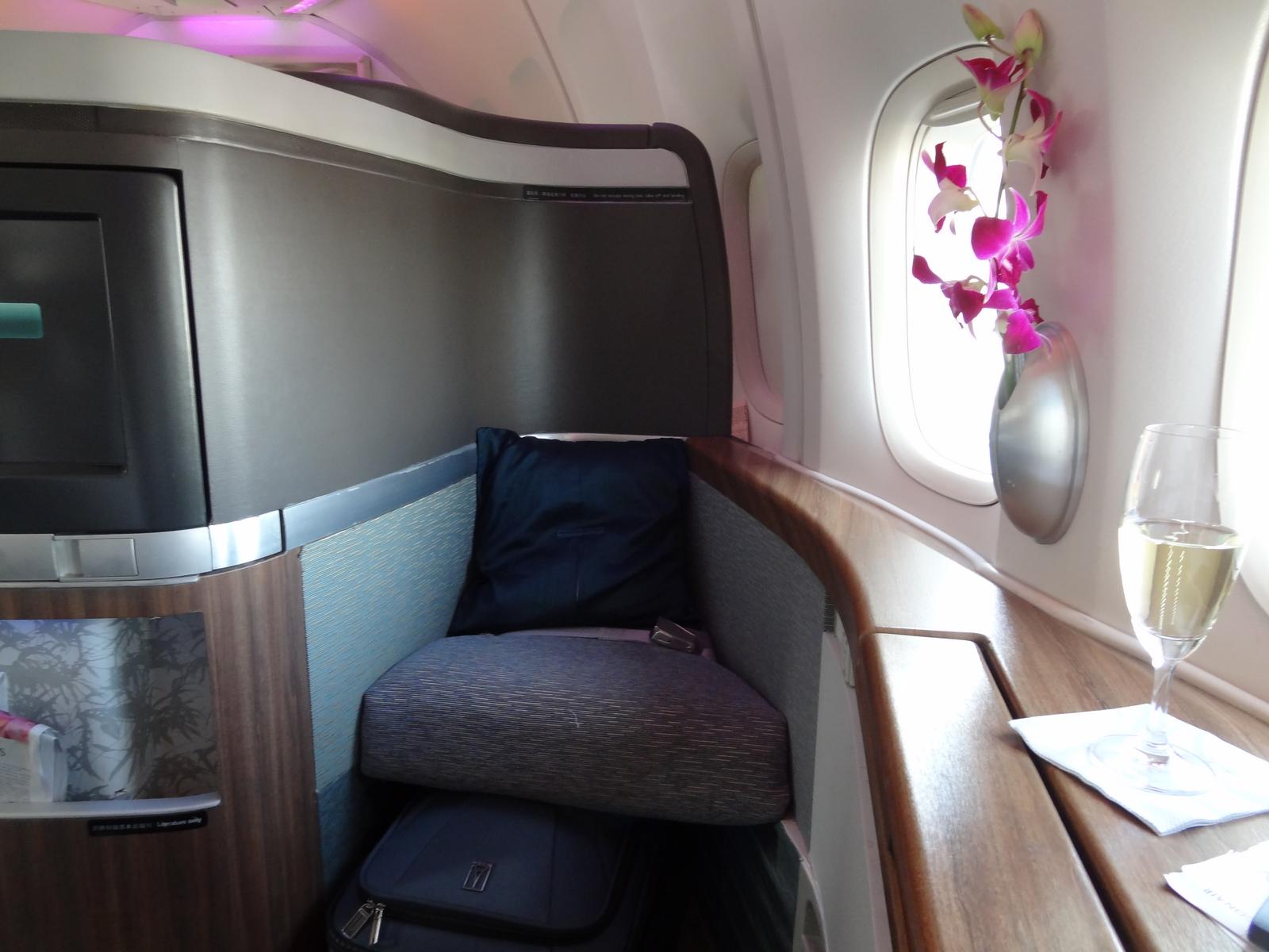 A Cathay Pacific 747 First Class cabin with a seat, window-mounted orchids, and a glass of champagne.