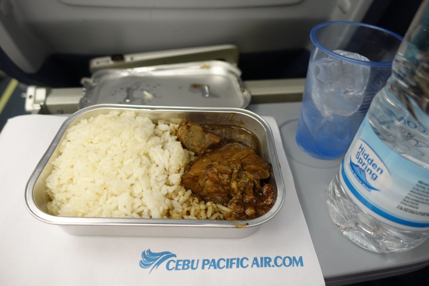 An airplane meal of rice and a meat dish, bottled water, and a cup of ice on a Cebu Pacific tray table.