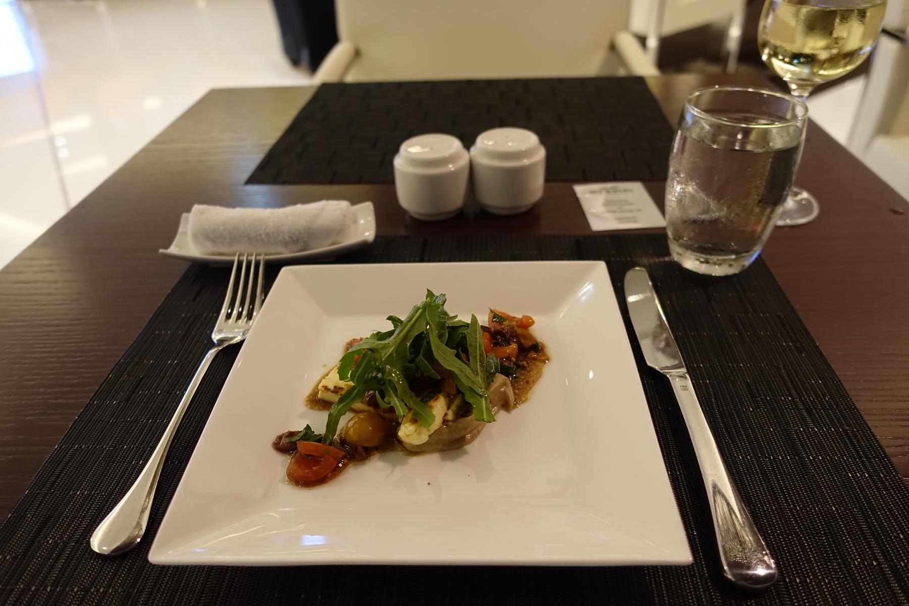 A beautifully plated meal of arugula, grilled cheese, and tomatoes, served on a white plate at a dark dining table with drinks.