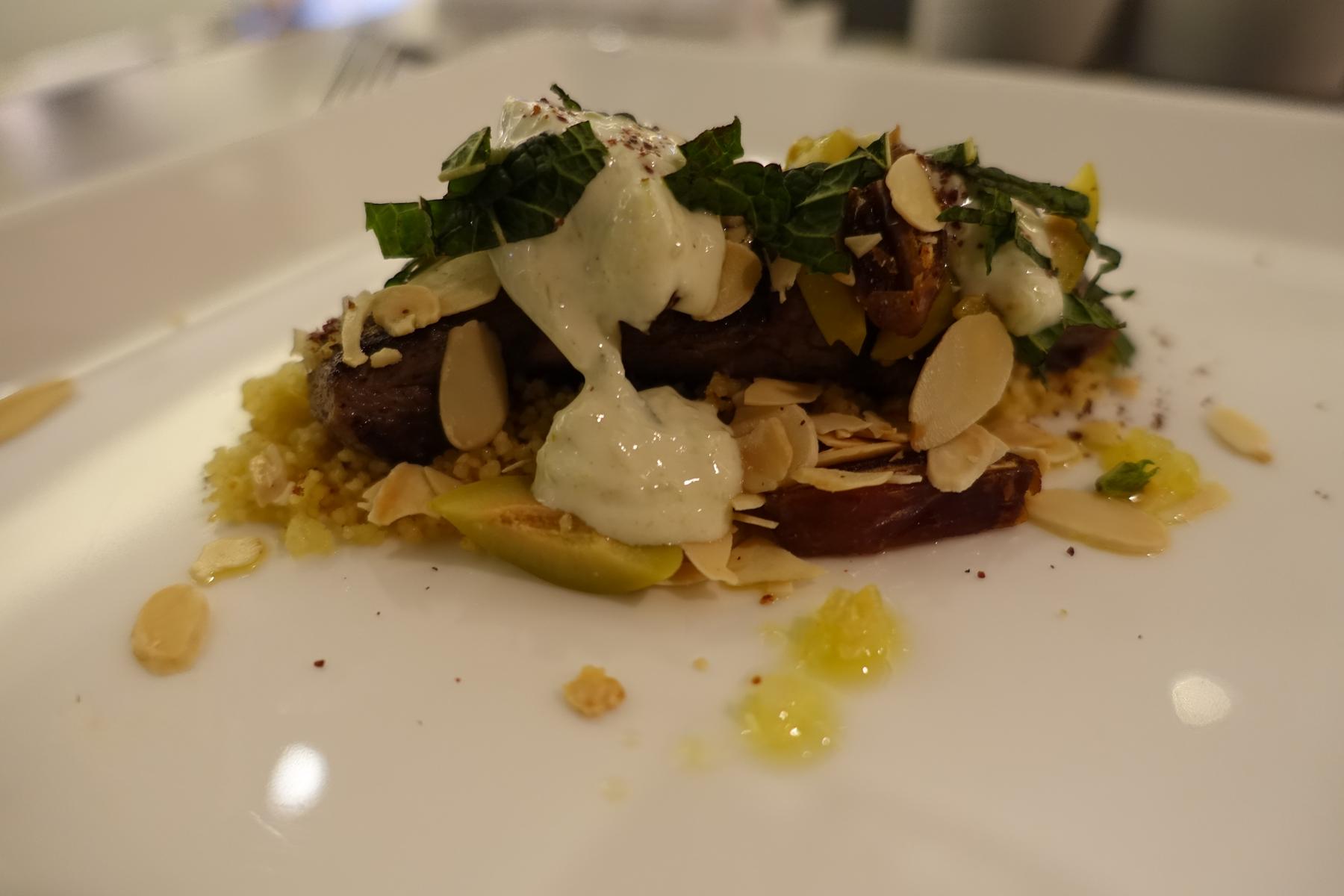 A plated dish of meat on couscous, topped with sliced almonds, green herbs, and a creamy white sauce.