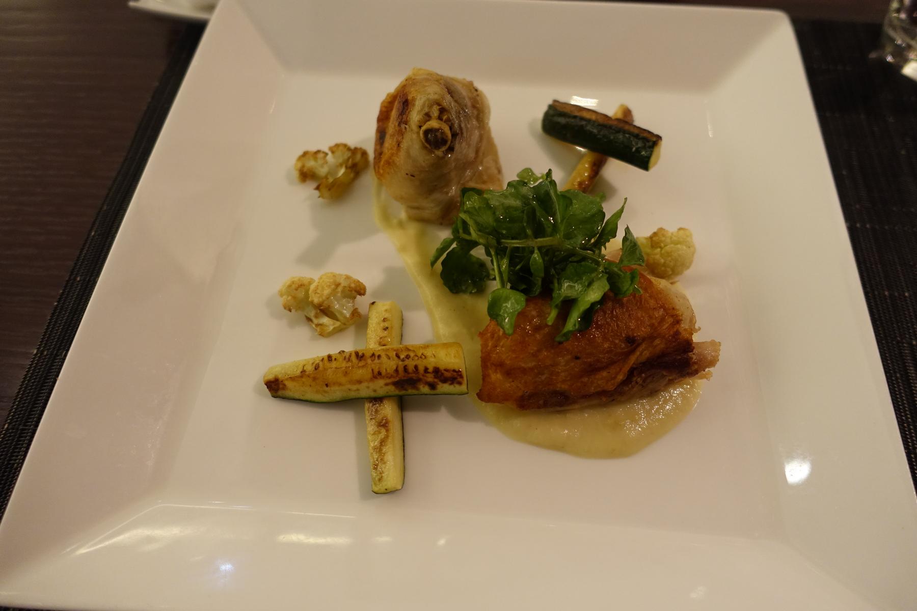 A gourmet plated meal featuring roasted poultry, grilled zucchini, cauliflower, and fresh greens.