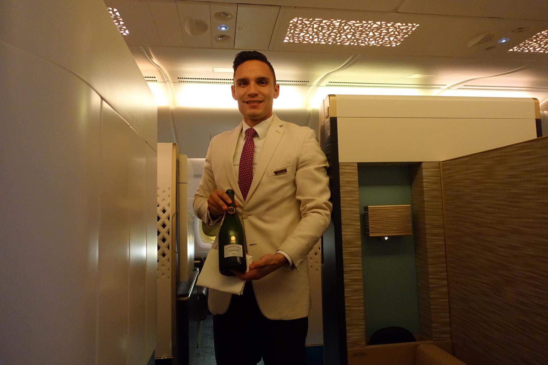 A smiling Etihad flight attendant in a light-colored suit holds a bottle of champagne in a luxurious airplane cabin.