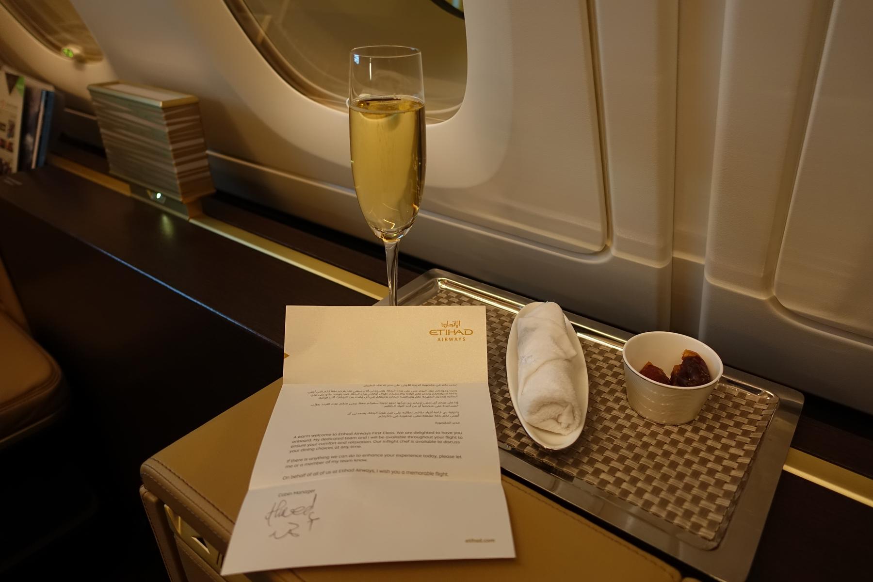 A welcome letter, champagne, warm towel, and dates are presented on a tray in an Etihad Airways First Class cabin.