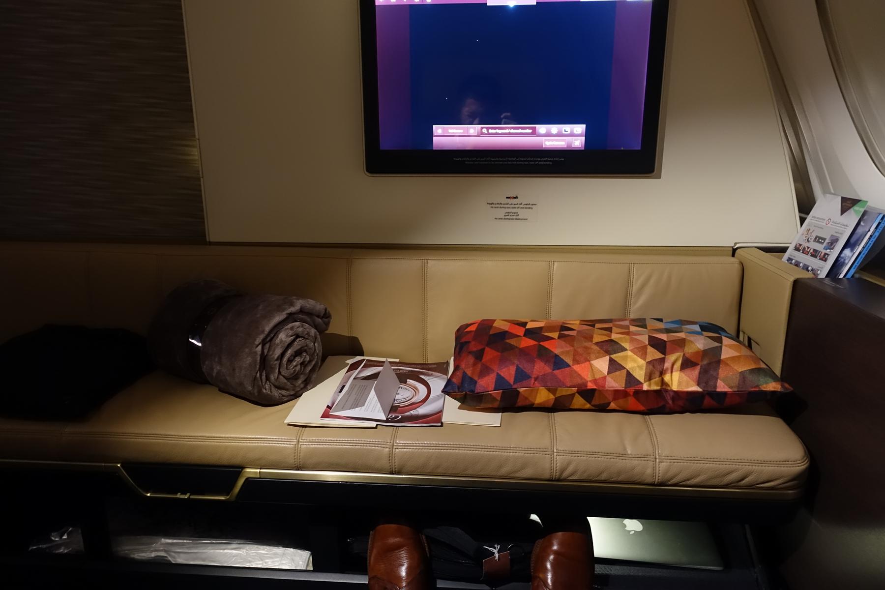 A luxurious Etihad A380 First Class apartment featuring a beige leather sofa, in-flight entertainment screen, blanket, and patterned pillow.