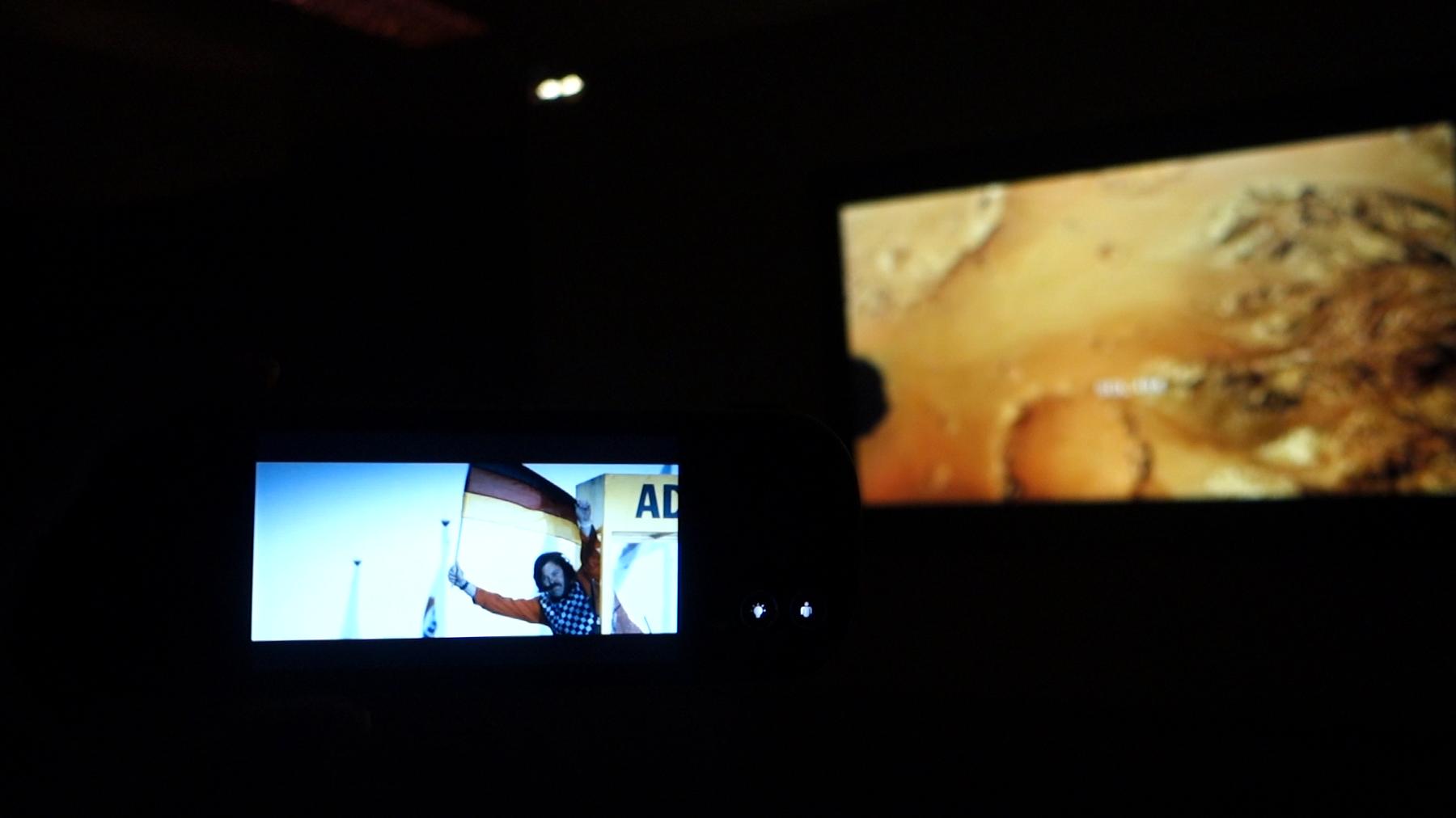 Two in-flight entertainment screens in a dark cabin show a man holding a German flag and a blurry orange landscape.