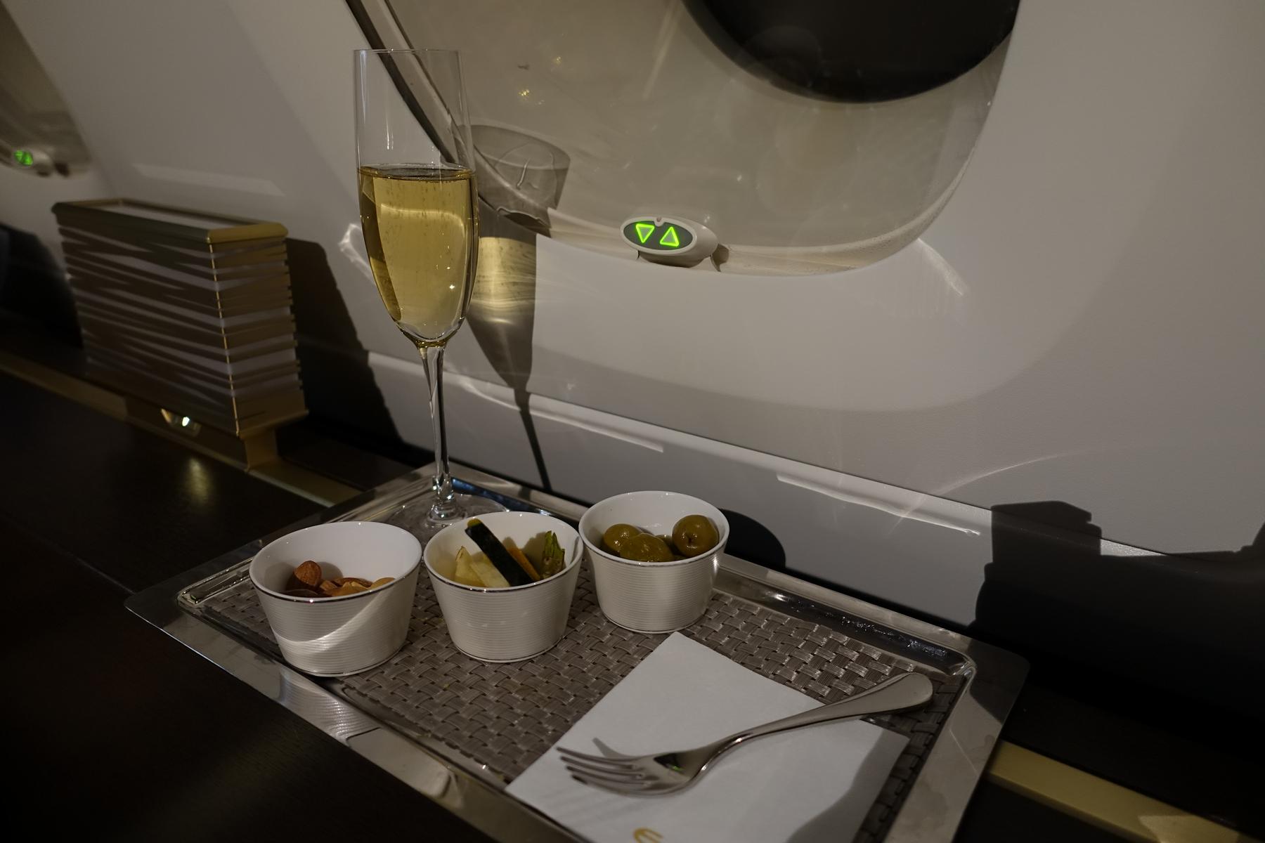 A flute of champagne and three snack bowls on a tray inside a luxurious airplane cabin.
