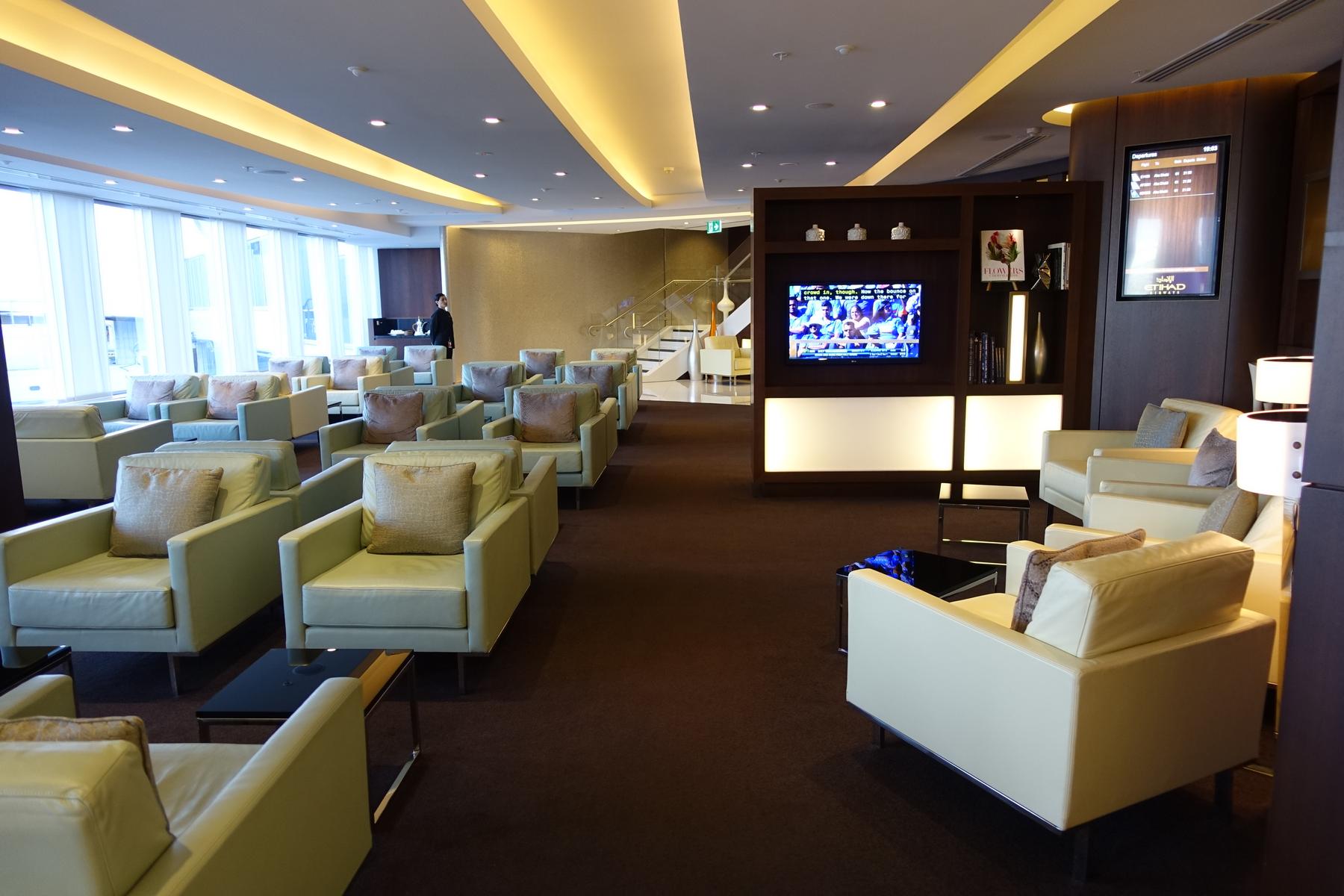 A luxurious Etihad First Class airport lounge with rows of comfortable armchairs, a large TV, and a departure board.