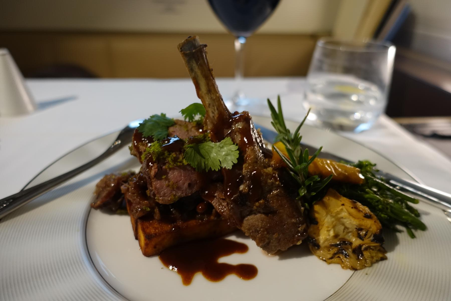 A perfectly plated rack of lamb with vegetables, sauce, and fresh herbs, served with drinks in a luxurious setting.