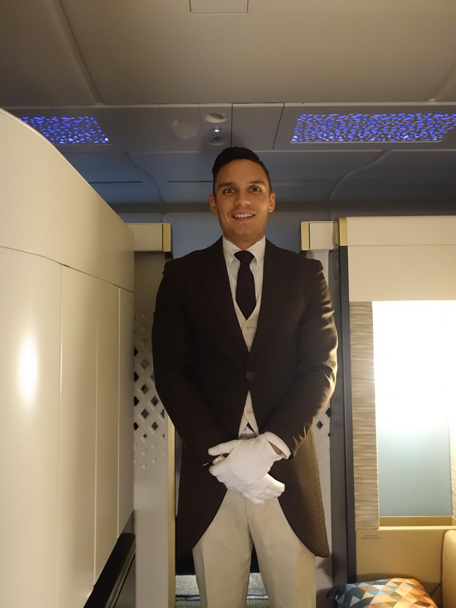 A smiling Etihad butler in a dark tailcoat and white gloves stands onboard an A380.