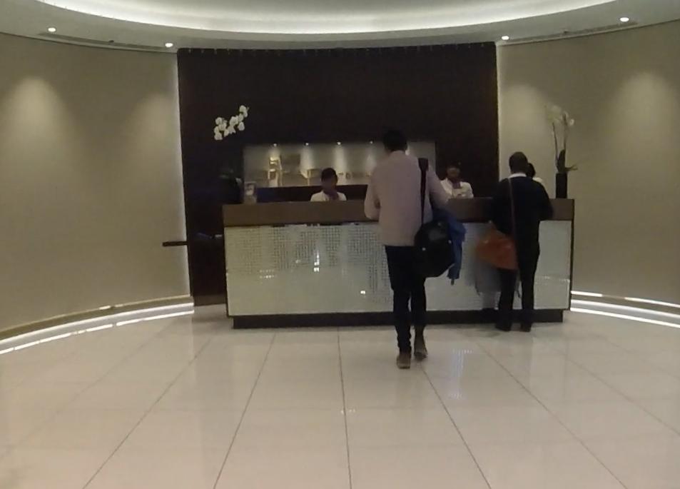 Travelers stand at a modern, brightly lit reception desk, likely in an airport business class lounge.