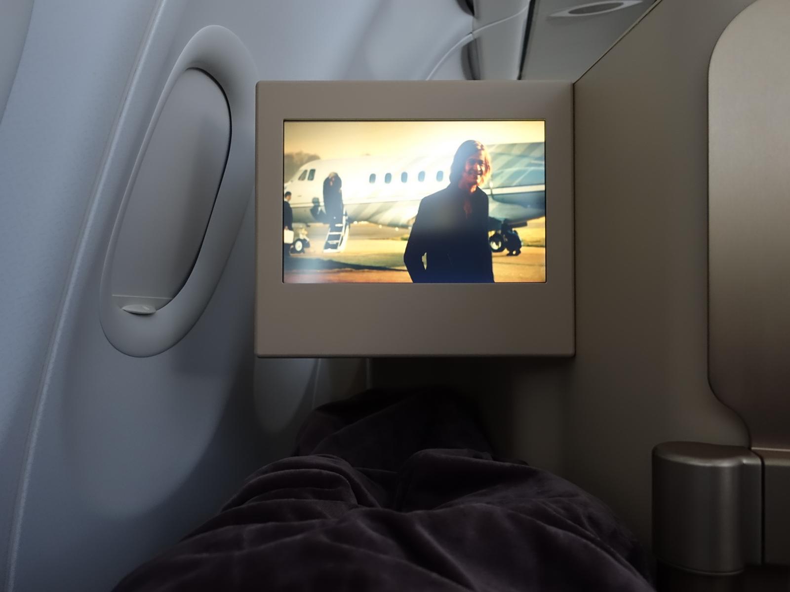 An Etihad Business Class seat showing a personal entertainment screen playing a scene with a man and a private jet, next to a window and a dark blanket.