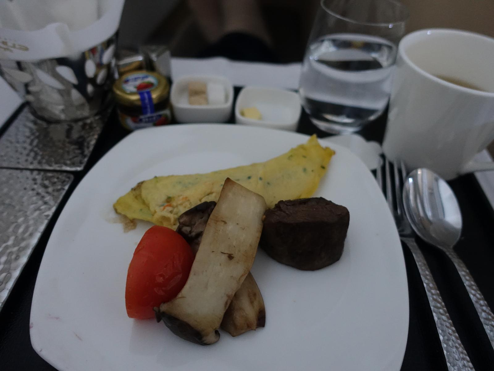 An Etihad Business Class meal on an airplane tray features an omelet, roasted meat, mushrooms, tomato, and beverages.