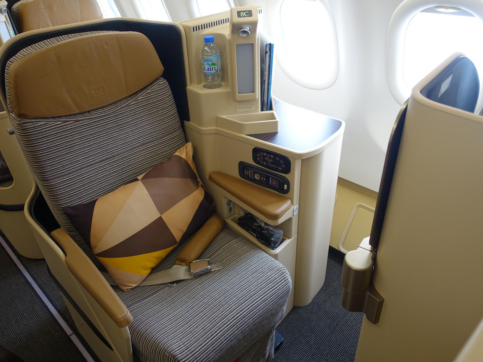 An Etihad Business Class airplane seat with a striped cushion, geometric pillow, and bottled water near a window.