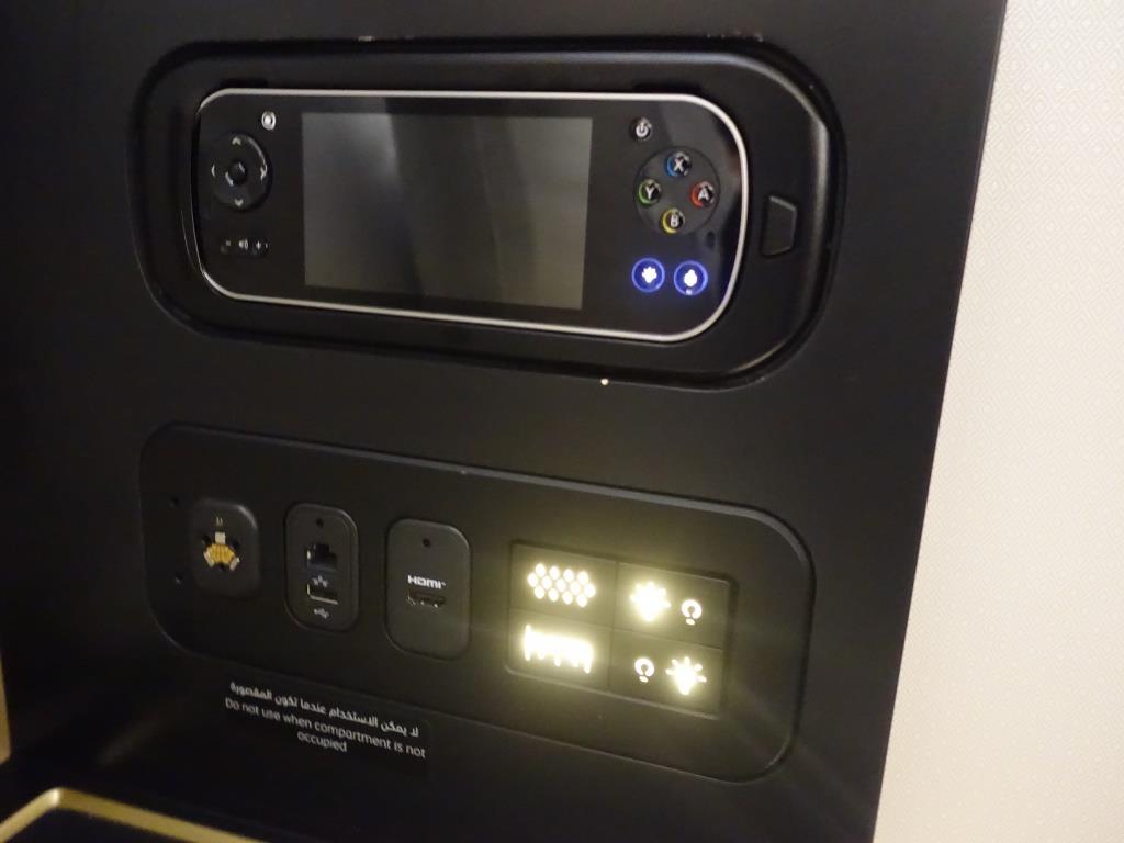 An airplane seat control panel features a handheld remote with a screen, power outlets, and illuminated comfort controls.