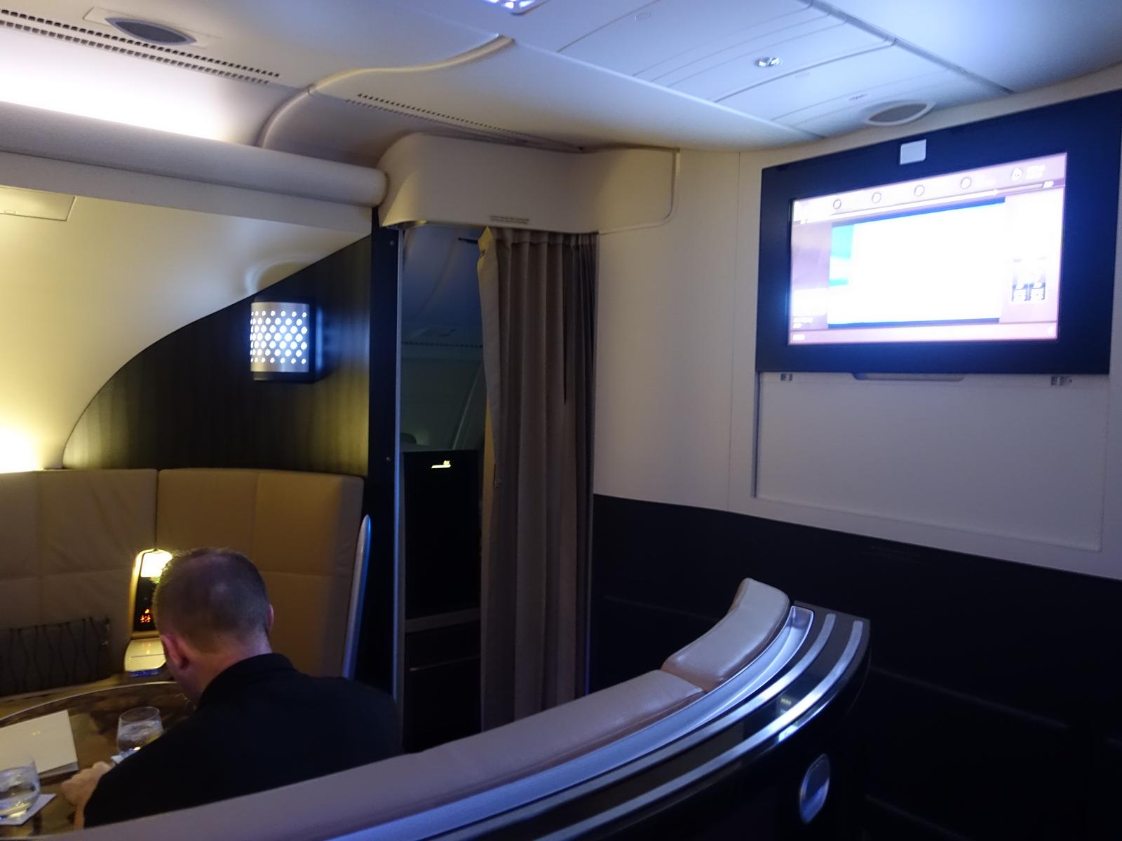 A man sits in a spacious Etihad A380 First Class apartment with a large entertainment screen on the wall.