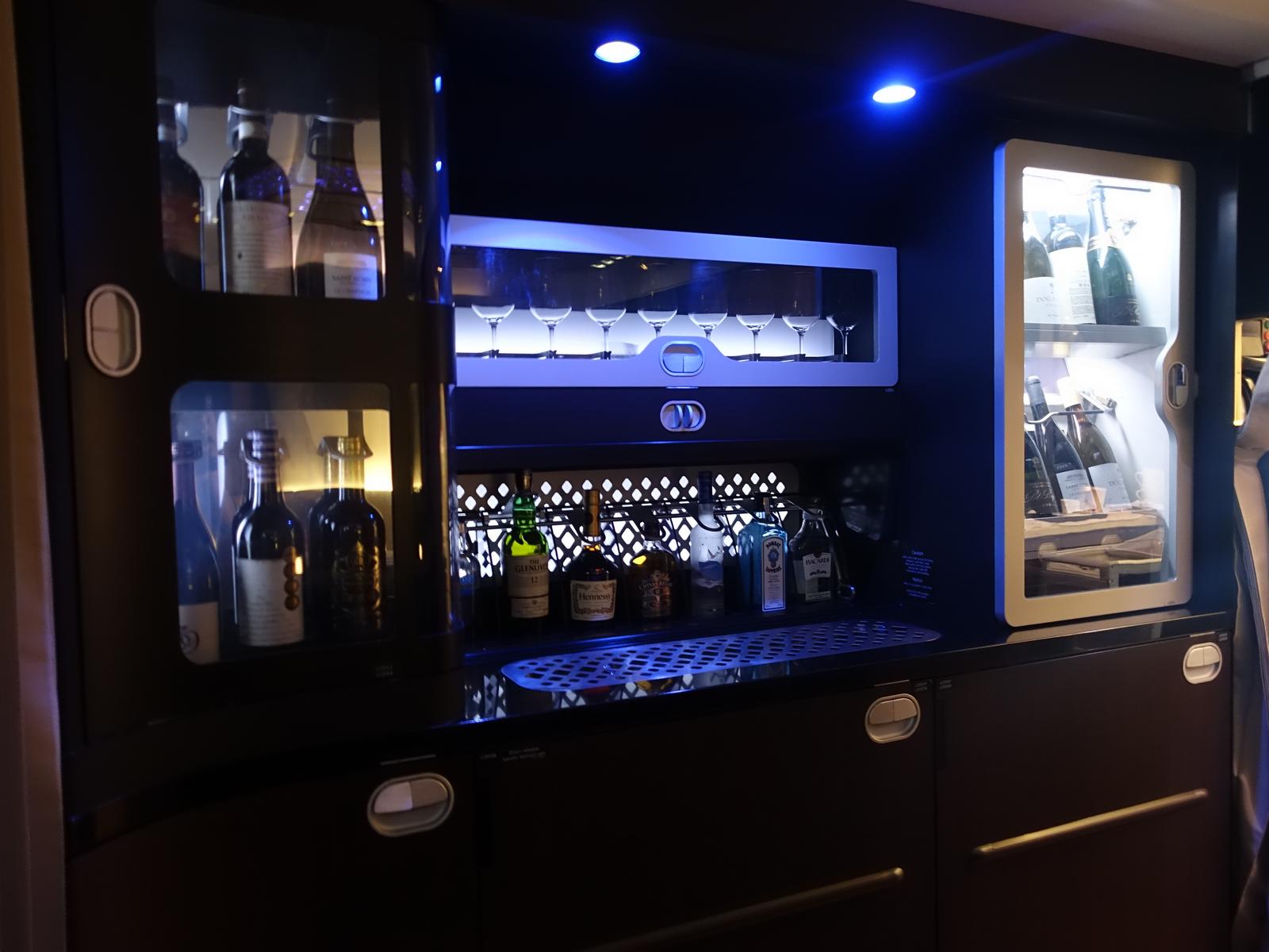 A luxurious bar area on an airplane features various liquor bottles, wine glasses, and a chilled wine fridge, all illuminated with blue accent lighting.