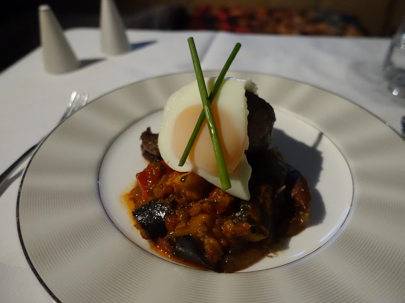 A gourmet dish of a soft-boiled egg with chives, served over a vegetable and meat medley on a white plate.