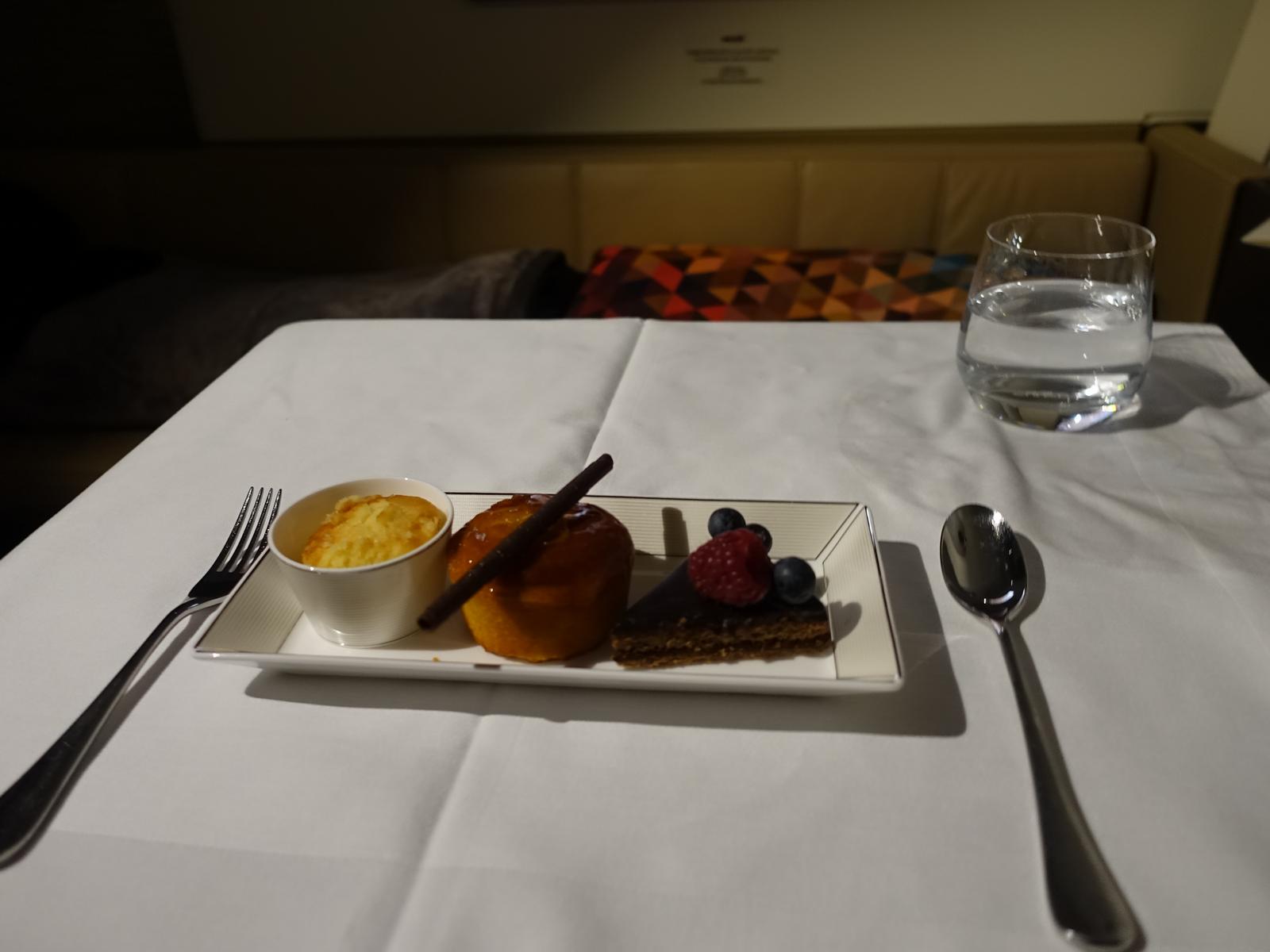 A luxurious dessert plate featuring chocolate cake, a glazed pastry, and a small dish of dessert, served on a white linen table in an Etihad First Class cabin.