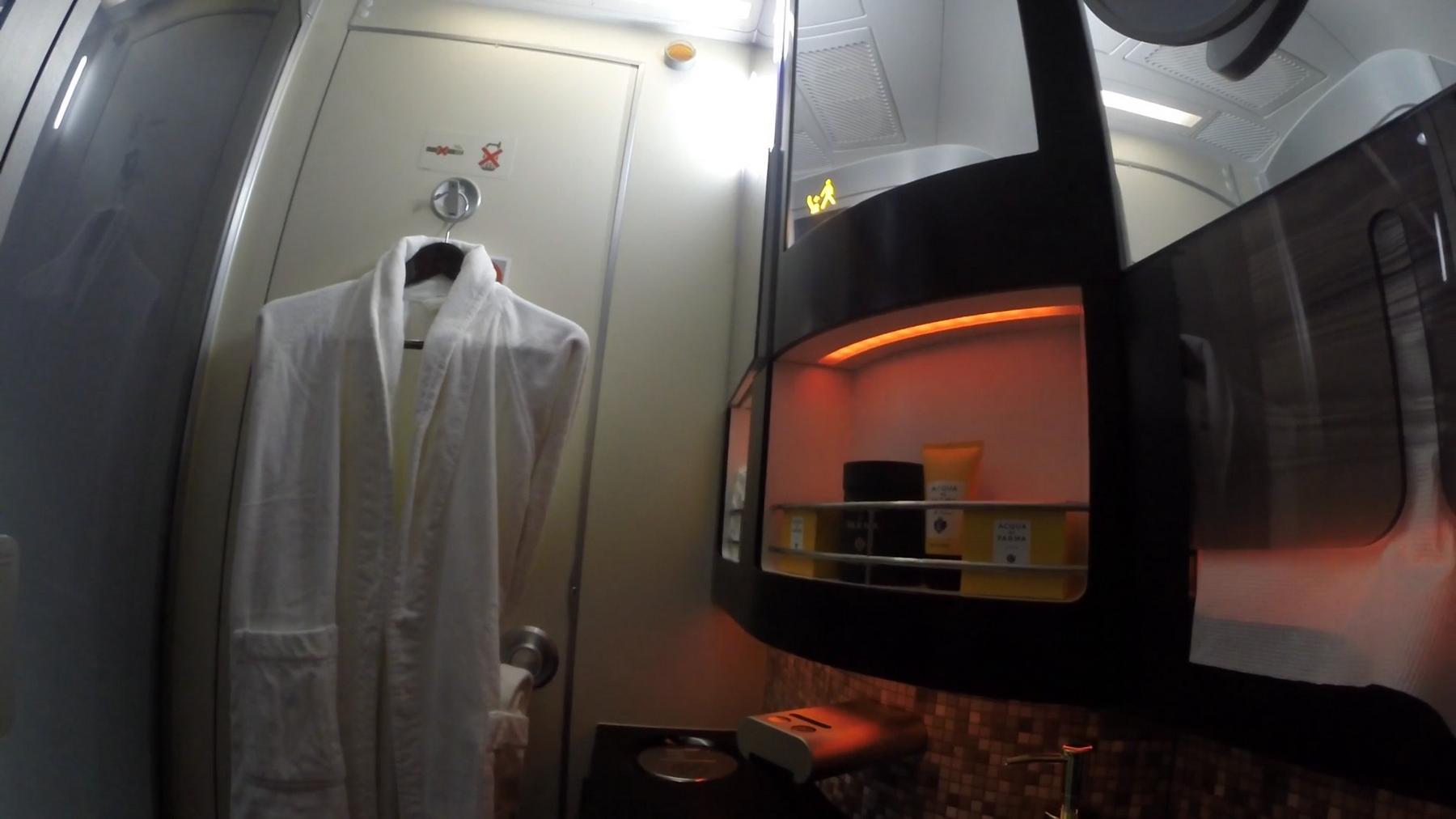 A white bathrobe hangs in a luxurious Etihad First Class lavatory, featuring an illuminated cabinet with premium toiletries.