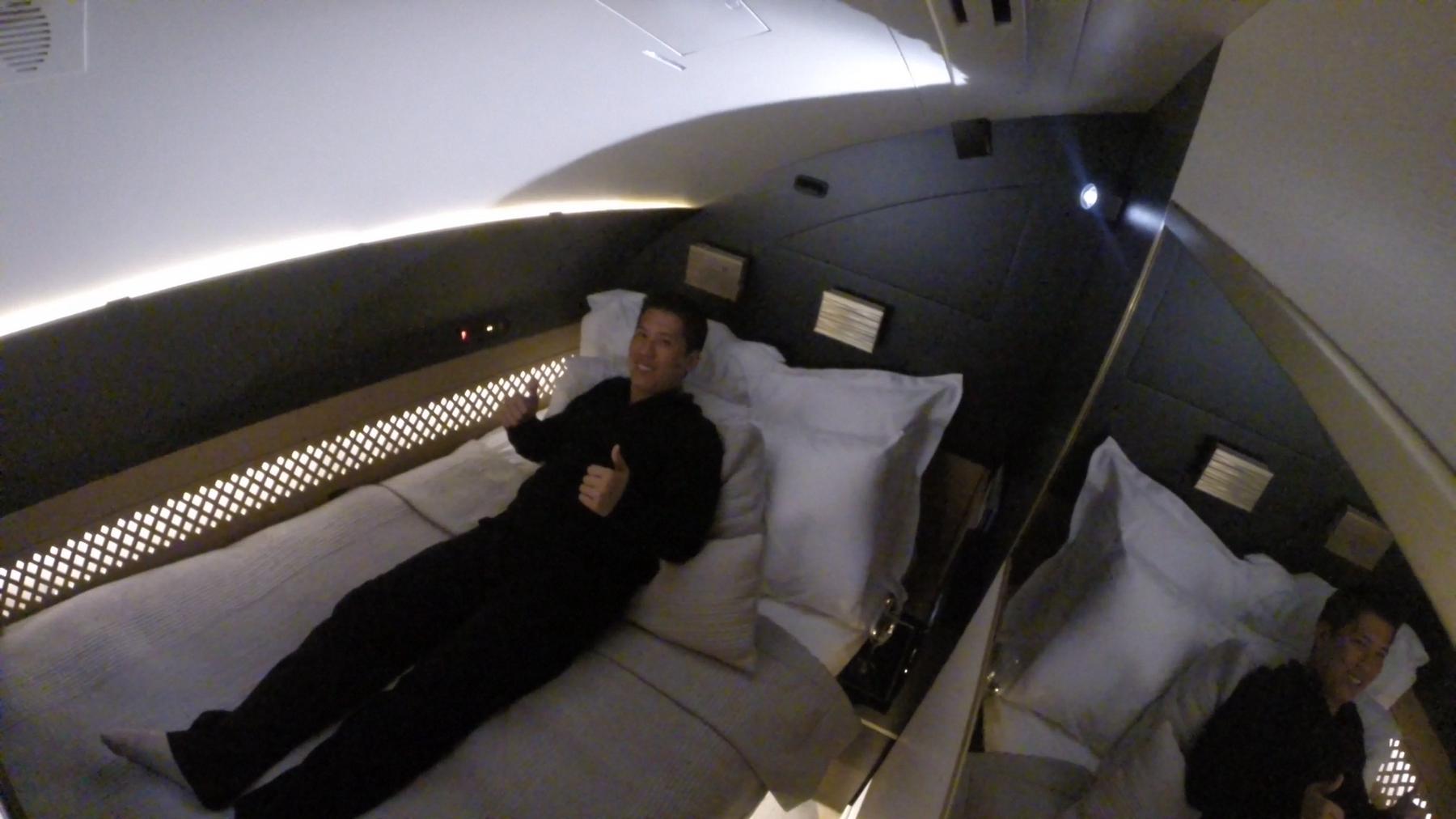 A man gives a double thumbs-up while lounging on a large bed in a luxurious airplane suite.