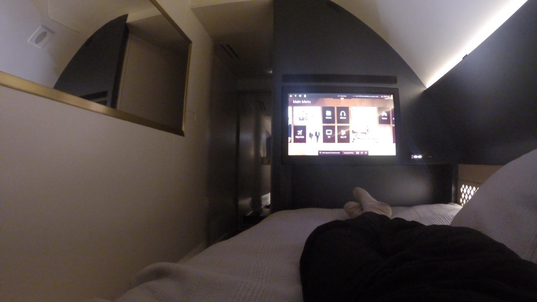 A first-person view of feet resting on a bed in a luxurious airplane suite, facing a large in-flight entertainment screen.