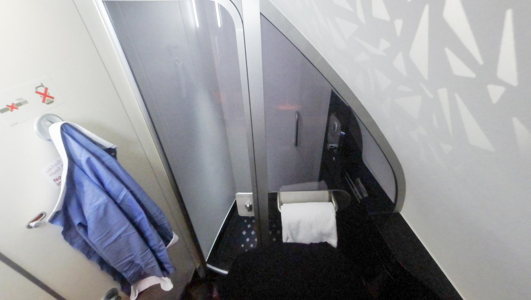 A luxurious Etihad A380 First Class lavatory featuring a blue robe on a hook, toilet paper, and modern grey and white decor.